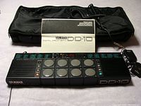 Full view of Yamaha DD-10 digital drum unit with power adapter, instruction manual, and black carrying case laid out on a surface.