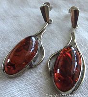 Close-up side view of both sterling silver amber earrings showing polishes oval amber stones with inclusions set in flowing silver frames with post backs.