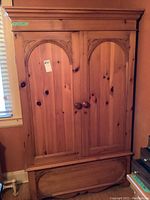 Front view of pine armoire showing two arched panel doors with wooden knobs and decorative hand carved accents at the top corners. Lower deep storage drawer visible with carved oval detail.