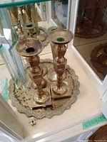 Two tall brass antique candle holders placed on a brass decorative tray in front of a mirrored background, showing tarnish and wear.