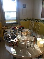 Wide view of a table filled with various mugs, bowls, glassware, silver plate items, and kitchen utensils in a yellow kitchen room.