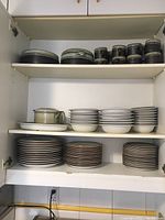 Photo shows multiple stacks of plates, bowls, cups, and saucers inside a cabinet shelf, including a teapot on the left side.