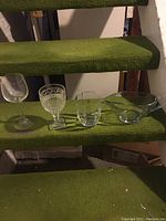 Four different types of glassware including a clear wine glass with stem, an ornate cut goblet, a plain tumbler, and a glass bowl placed on green carpeted stairs.