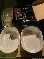 Two white glass baking dishes and a boxed metal wine opener set including wing corkscrew and bottle stoppers on a wooden base.