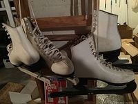 Two pairs of white ladies figure skates displayed on a wooden chair, one pair showing a size around 7 and the other around 8, with black skate guards on the blades.