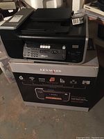 Black Lexmark X6675 Pinnacle Professional Series printer sitting on top of its original box on a concrete floor, surrounded by other items.