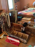 Wooden necklace tree standing next to a mirror with various necklaces hanging, showing beads, faux pearls, and chains on wooden stand; basket and brushes in foreground.