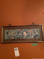 Framed silk textile art hanging on orange wall showing detailed embroidered butterfly and flowers, gold bamboo-style frame, dimensions 9x21 inches.
