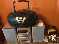Photo showing Sony speakers, RCA boombox, WestClox clock, iPod with earphones, and remote control on a shelf against an orange wall.