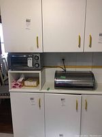 Overall view showing the toaster oven on a shelf beside a metal bread box on the counter below white cabinets with yellow handles.