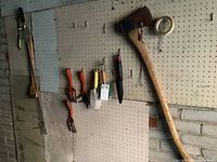 Image shows multiple hand tools hanging on a pegboard including a hatchet, garden forks, trowel, gardening knife and a handheld electric trimmer by Black & Decker.