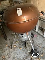 Side view of copper-toned round kettle barbecue resting on metal tripod legs with two wheels visible. Shows wear and some debris on ground.