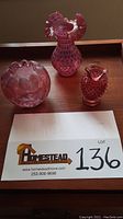 Three cranberry glass pieces displayed together on a wooden surface including a ruffled edge bowl, hobnail pitcher, and larger hobnail vase