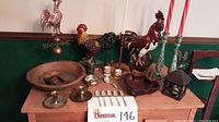 Wide shot of collection on wooden table showing brass and copper rooster weathervane, cast iron rooster doorstop, wooden bowls with chopping tool, brass candlesticks, silver spoons and ladle, pewter cup, brass snuff container, and small lantern.