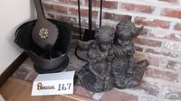 Metal ash bucket, fireplace tool set on stand, bellows inside ash bucket, and resin cherubs statue near brick wall.