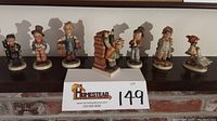 Wide view of all 7 Hummel figurines lined on a shelf including boy with tools and girl with goose