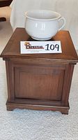 View of the wooden chamber pot cabinet topped with the white porcelain chamber pot, showing front panel and part of the top.