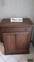 Front view of wooden hamper cabinet showing drawer, two doors, and decorative handles.