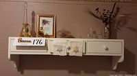 Front view of Lochner Woodworking white painted wall shelf with three drawers. Shelf includes vintage glass bottle, framed art piece, small covered glass dish, hanging embroidered cloths, and a dry floral arrangement in vase as displayed.