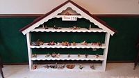 Front view of white wooden chalet display with scalloped edging and four shelves filled with assorted handmade dolls.