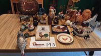 Full view of the collection displayed on the table showing various rooster and chicken figurines, a decorative plate, wooden plaques, and a clear glass bowl with black rooster designs on a metal stand.