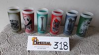 Six souvenir glass tumblers standing in a row on a lace cloth, showcasing colorful fair-themed images from the 1962 Seattle World's Fair.