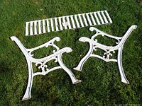 All bench parts laid on grass showing two side frames and three slatted seat sections, painted white, detailed ornate scroll and leaf decoration on frames.
