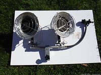 Top-down photo showing two circular metal propane burners side by side, mounted on a white board on grass background.