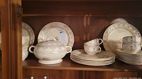 Wooden cabinet shelf holding the china set including plates, covered bowl, and stacked teacups.