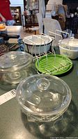 Overview photo showing clear glass Corningware pots and casseroles with lids, metal wire rack, and green glass plate on a kitchen counter.