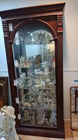 Full view of dark wood framed curio cabinet showing glass front and sides, five glass shelves containing display figurines, and arched glass door.
