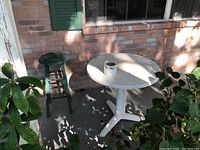 Plastic white round patio table with tripod base beside green wooden bar stool, outdoor setting on concrete porch, shadows visible.