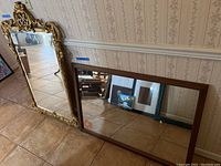 Photo showing both mirrors leaning against a wall on tiled floor; ornate gold tone framed mirror on left, simple wood framed mirror on right.
