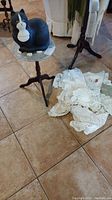 View of small marble-top table holding ceramic cat cookie jar, with lace doilies scattered on floor beside it