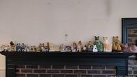 Mantle shelf displaying 19 ceramic cat figurines and a large painted wooden cat plaque, varied sizes and colors.