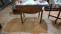 Front angled view of the vintage wooden drop leaf table with lace cloth on top and visible scratches on surface.