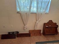 Five vintage wooden wall shelves arranged against a beige wall under two small windows with curtains. Shelves vary in size, shape, and wood tone.