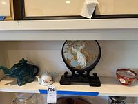 Shelf with teal elephant teapot, small white porcelain teapot, embroidered art plate on stand, and red decorative bowl