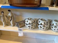 Three white spherical ceramic candle holders with symmetrical cut-outs and black metal handles alongside three Asian-style ceramic figurines on a shelf.