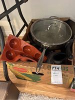 Photo showing a stainless steel pan with glass lid, silicone baking mold, and black plastic food processor parts inside a cardboard box.