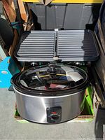 Overall photo showing a stainless steel Cook's Essentials slow cooker with glass lid and front dial control, sitting below a Hilton Beach electric griddle with ribbed cooking surface.