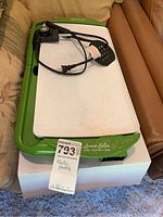 Electric griddle with white cooking surface, green base, temperature dial, and cord coiled on top, placed on cardboard box