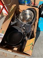 Top view of box showing non-stick frying pan, stainless steel bowls, and various black cooking utensils including spatulas.