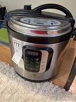 Photo showing the full side and top view of a Cook's Essentials electric pressure cooker sitting on carpeted floor, featuring stainless steel body and black plastic handle.