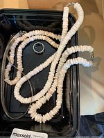 Photo shows two white puka shell necklaces with numerous small shell beads placed in a tray, along with a koa and abalone men’s ring size 9.