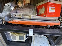 Ridgid tile saw with orange safety guard on top of black plastic shelving. Small white window air conditioner inside shelving below saw. Box of Commercial Electric Color Changing Recessed Trim visible to the right.