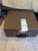 Closed vintage typewriter case sitting on a carpet, dark brown textured surface with a black handle and metal latch.