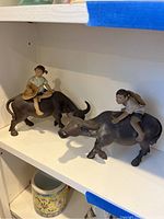 Two metal figurines of children riding water buffalo on a white shelf.