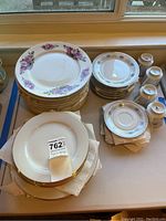 Photo showing stacks of floral patterned plates including sets with purple floral designs and pink flower clusters edges, along with 4 porcelain tea cups and saucers.