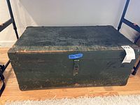 Front view of the vintage wooden chest showing full rectangular shape, dark green paint with wear, metal corner protection and central latch.